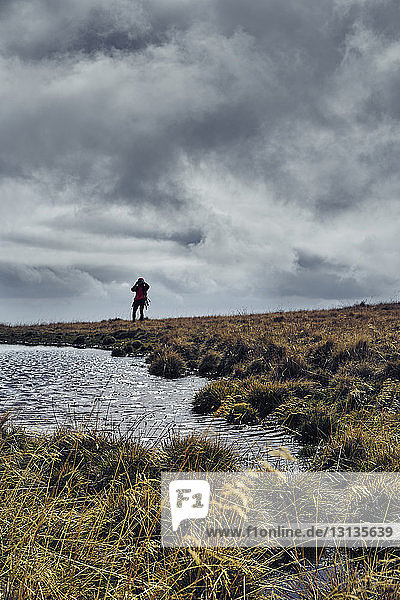 Mid distance view of man standing on grassy field by lake against cloudy sky