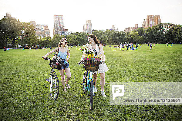 Smiling lesbian couple walking with bicycles on grassy field at park