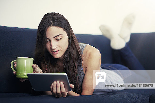 Junge Frau hält Kaffeetasse und benutzt digitales Tablett auf dem Sofa