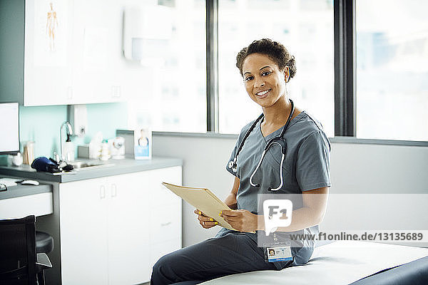 Porträt einer glücklichen Ärztin  die eine Akte hält  während sie in der Klinik auf dem Untersuchungstisch sitzt