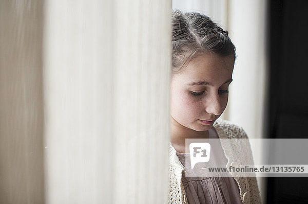 Trauriges Mädchen durch Fenstervorhänge zu Hause