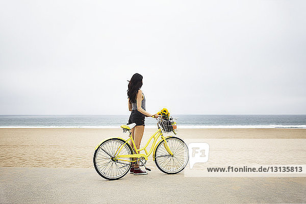 Frau steht mit Fahrrad am Strand vor klarem Himmel
