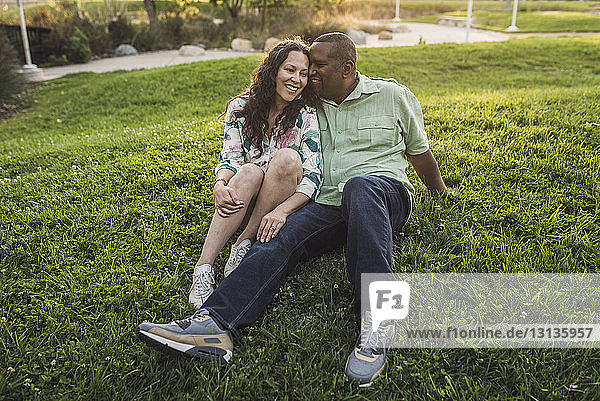 Das glückliche Paar unterhält sich in voller Länge  während es auf einem Grasfeld im Park sitzt