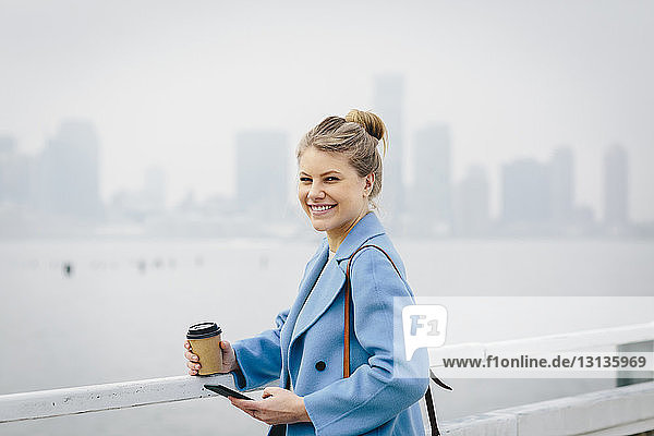 Portrait of cheerful young woman holding disposable cup and smart phone while standing by river in city