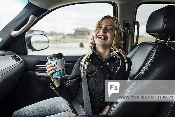Cheerful girl holding disposable cup while sitting in car
