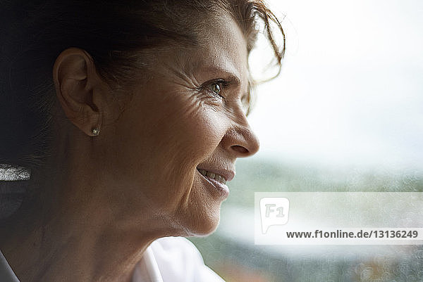 Nahaufnahme einer lächelnden Frau  die durch ein nasses Fenster schaut  während sie zu Hause steht