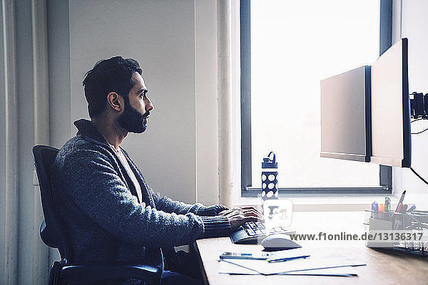 Seitenansicht eines Geschäftsmannes  der einen Desktop-Computer benutzt  während er im Büro am Schreibtisch sitzt