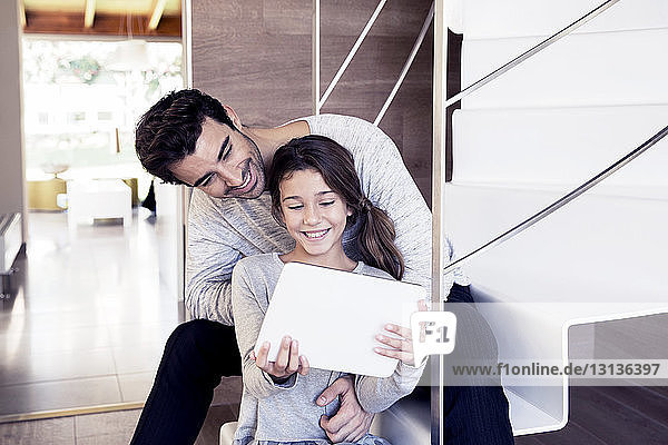 Lächelnder Vater und Tochter schauen auf das digitale Tablett  während sie auf Stufen sitzen