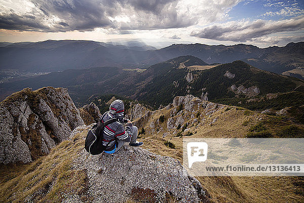 Rückansicht eines männlichen Rucksacktouristen  der auf einem Berg vor bewölktem Himmel sitzt