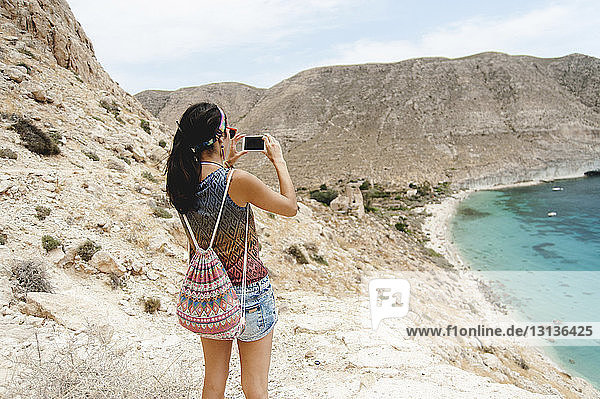 Rückansicht einer Frau mit Rucksack  die mit einem Smartphone das Meer fotografiert