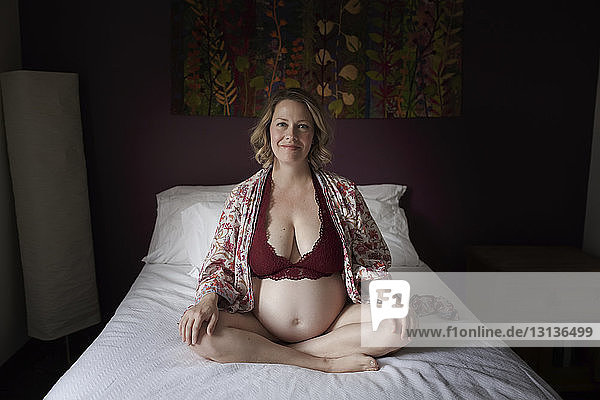 Porträt einer schwangeren Frau in Dessous  die zu Hause auf dem Bett sitzt