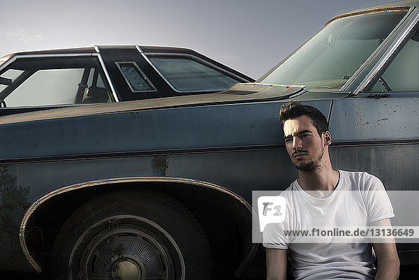 Thoughtful young man looking away while sitting against old vintage car