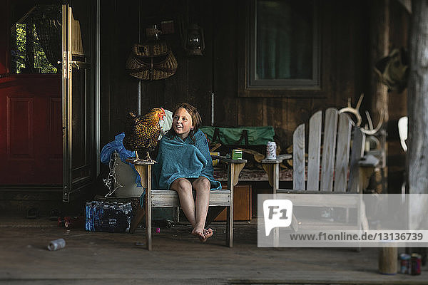 Girl wrapped in blanket looking at hen while sitting on porch