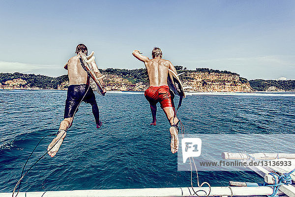 Rückansicht von hemdlosen männlichen Freunden mit Surfbrettern  die vor blauem Himmel ins Meer springen