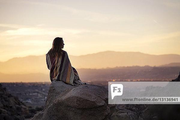 Rückansicht einer in eine Decke gehüllten Frau  die bei Sonnenuntergang auf einem Felsen gegen den Himmel sitzt