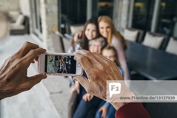 Abgetrennte Hände eines älteren Mannes fotografieren Familie durch ein Smartphone auf der Veranda