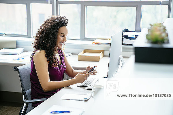 Smiling businesswoman using mobile phone while sitting at desk in office