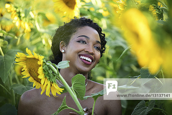 Nahaufnahme einer glücklichen Braut  die wegschaut  während sie inmitten von Sonnenblumen auf einem Bauernhof steht