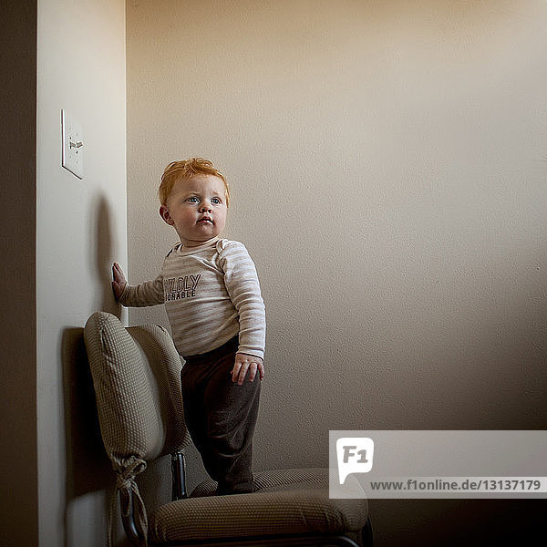 Cute baby boy looking away while standing on chair against wall at home