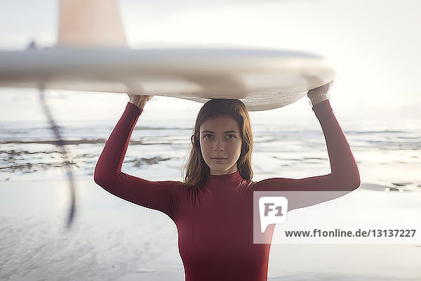Porträt einer jungen Frau  die am Strand stehend ein Surfbrett auf dem Kopf trägt
