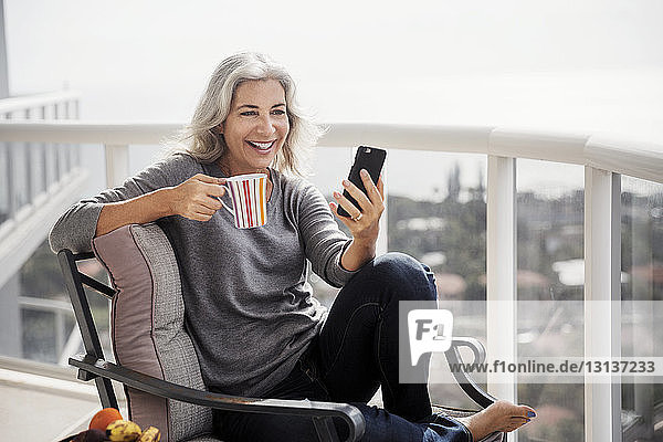 Glückliche reife Frau benutzt Smartphone beim Kaffee trinken auf dem Balkon