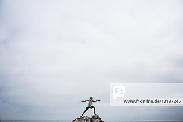 Frau übt die Pose von Worrier 2 auf Felsen im Meer vor bewölktem Himmel