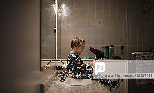 Seitenansicht eines Jungen  der einen Haartrockner hält  während er zu Hause am Badezimmer-Waschbecken sitzt