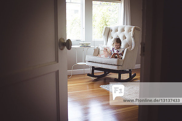 Mädchen liest Bilderbuch  während sie zu Hause auf einem Schaukelstuhl sitzt und durch die Tür gesehen wird