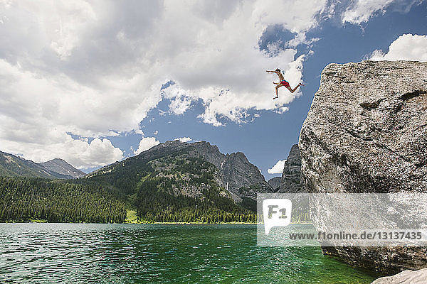 Low angle view of teenager cliff jumping in lake against cloudy sky