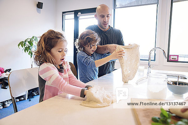Father assisting son and daughter in preparing dough at kitchen table