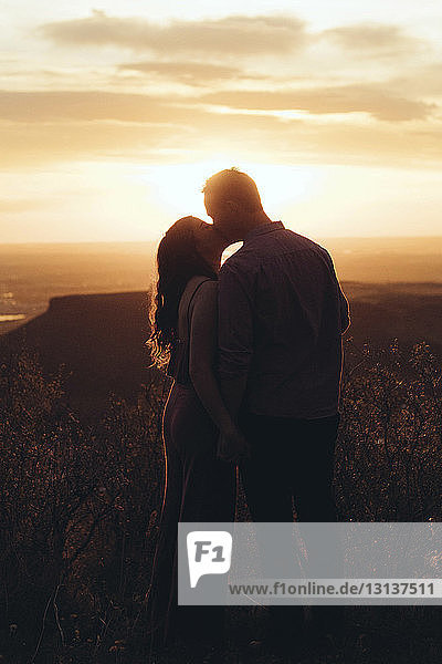 Junges Paar küsst sich  während es bei Sonnenuntergang auf dem Feld gegen den Himmel steht