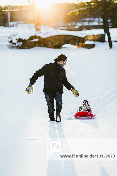 Father pulling sled with daughter on snow covered field at park