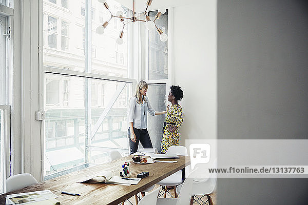 Seitenansicht von Geschäftsfrauen  die sich unterhalten  während sie am Fenster im Sitzungssaal stehen