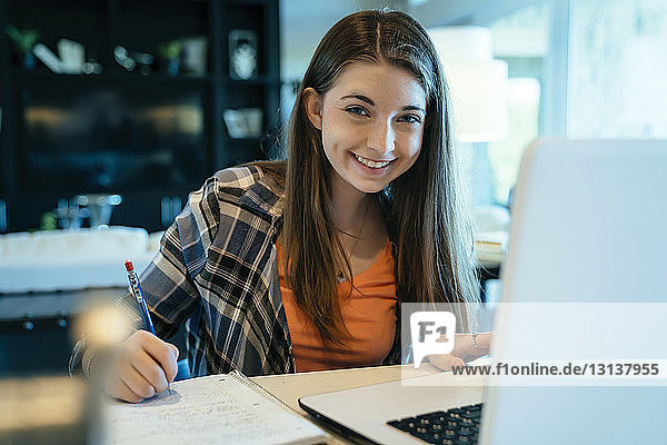 Porträt einer glücklichen Teenagerin  die zu Hause am Laptop Hausaufgaben macht