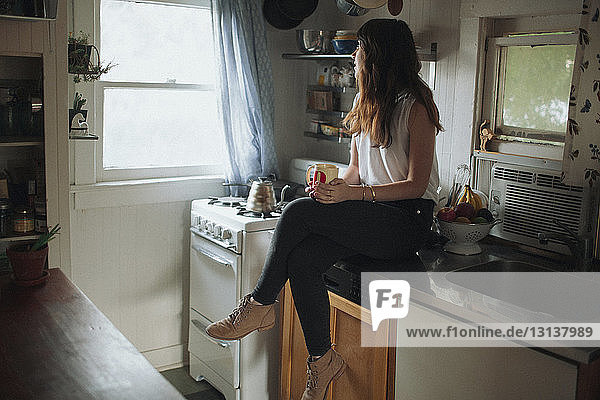 Nachdenkliche Frau hält Kaffeetasse  während sie auf der Küchentheke sitzt