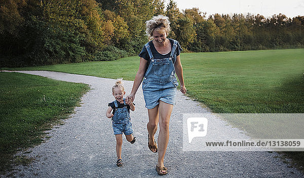 Cheerful mother and daughter wearing bib overalls while running on dirt road