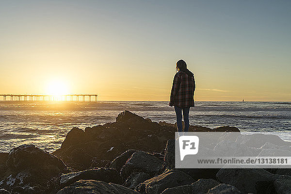 Side view of woman standing on rocky shore against sky during sunset