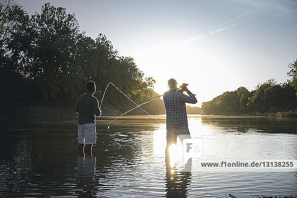 Rear view of male friends fishing while standing in lake against sky