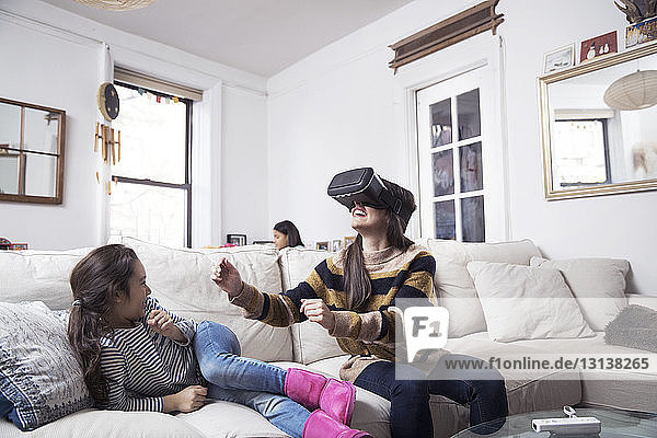 Daughter looking at mother enjoying virtual reality simulator while sitting on sofa