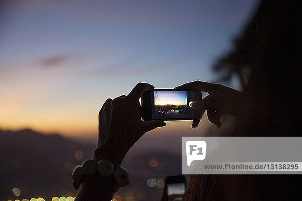 Frau fotografiert beleuchtete Stadtlandschaft mit Smartphone bei Sonnenuntergang