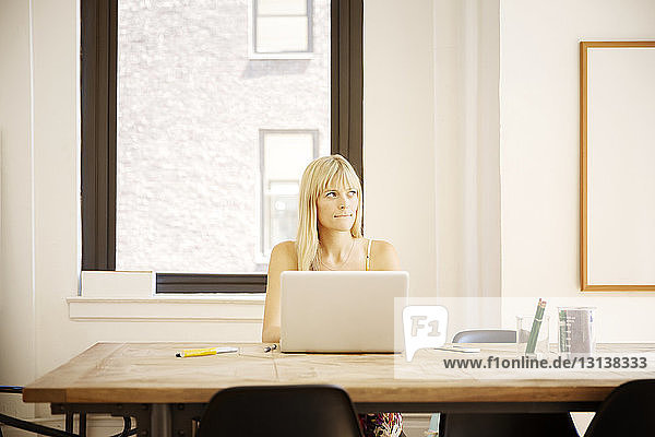 Nachdenkliche Geschäftsfrau sitzt mit Laptop am Schreibtisch im Kreativbüro