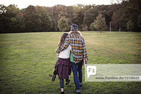 Rear view couple carrying skateboards while walking on grassy field