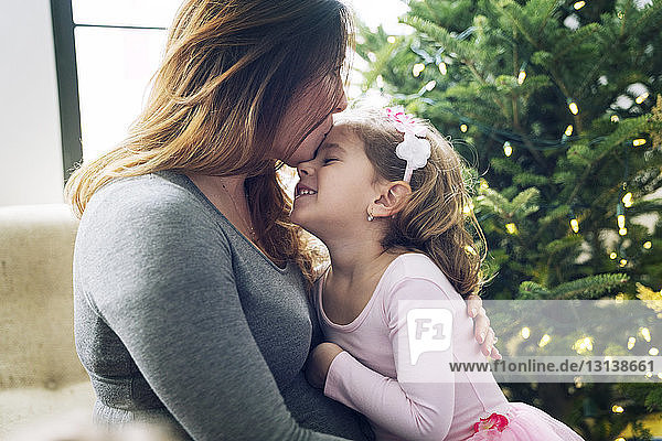 Nahaufnahme einer Mutter  die zu Hause ihre Tochter am Weihnachtsbaum küsst