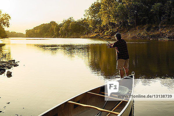 Rear view of mid adult man fishing while standing by boat in lake during sunset