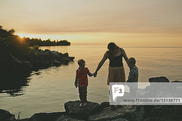 Mother with sons standing on rocks at lakeshore during sunset