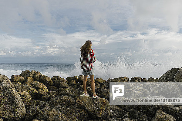 Junge Frau in voller Länge schaut aufs Meer  während sie auf einem Felsen vor bewölktem Himmel steht