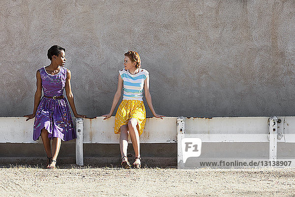 Multi-ethnische Freundinnen unterhalten sich  während sie auf einem Geländer an der Wand sitzen
