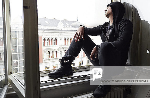 Junger Mann in schwarzer Kapuzenjacke in voller Länge  der zu Hause auf dem Fensterbrett sitzt