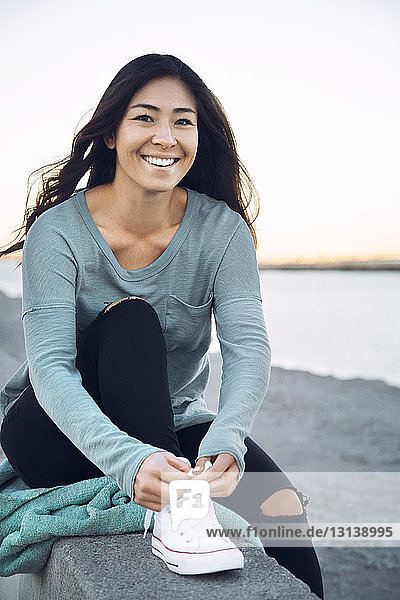 Porträt einer glücklichen Frau  die Schnürsenkel bindet  während sie auf einer Stützmauer am Strand sitzt