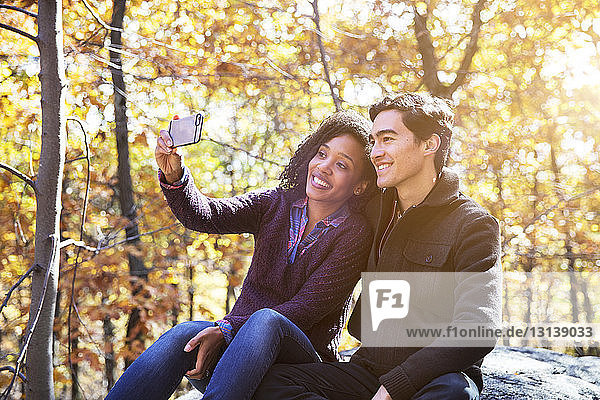Woman taking selfie while sitting with boyfriend on rock in forest